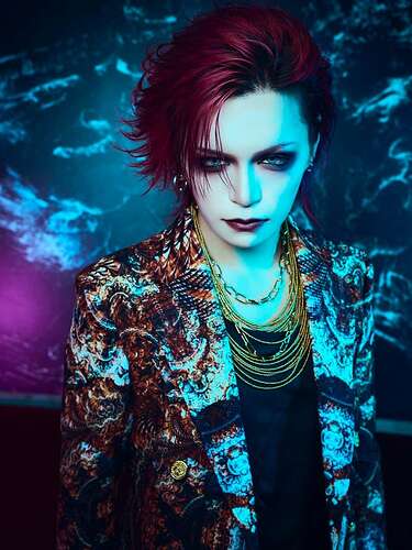 Dark red hair, dark vibe eye make-up and lipstick, black shirt with intricate patterned blazer in white, brown and golden shades, golden necklaces.