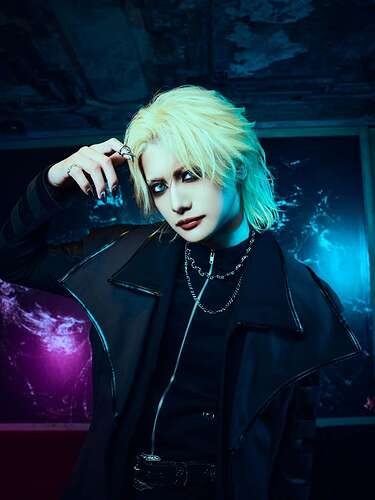 Light blond chin long hair, shorter at the sides, dark vibe eye make-up and lipstick, black shirt with a zipper, over it a wider coat like thing in black, two different silver necklaces, one claw like ring at the tip of an index finger.