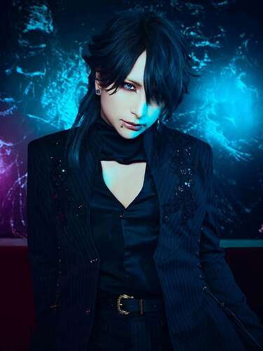 Black hair with some slight blue shade, dark vib eye make-up and lipstick, black suit vest like top with some addition fabric around the neck, black blazer jacket with sequins and some kinda lace adornment coming down from the shoulders.